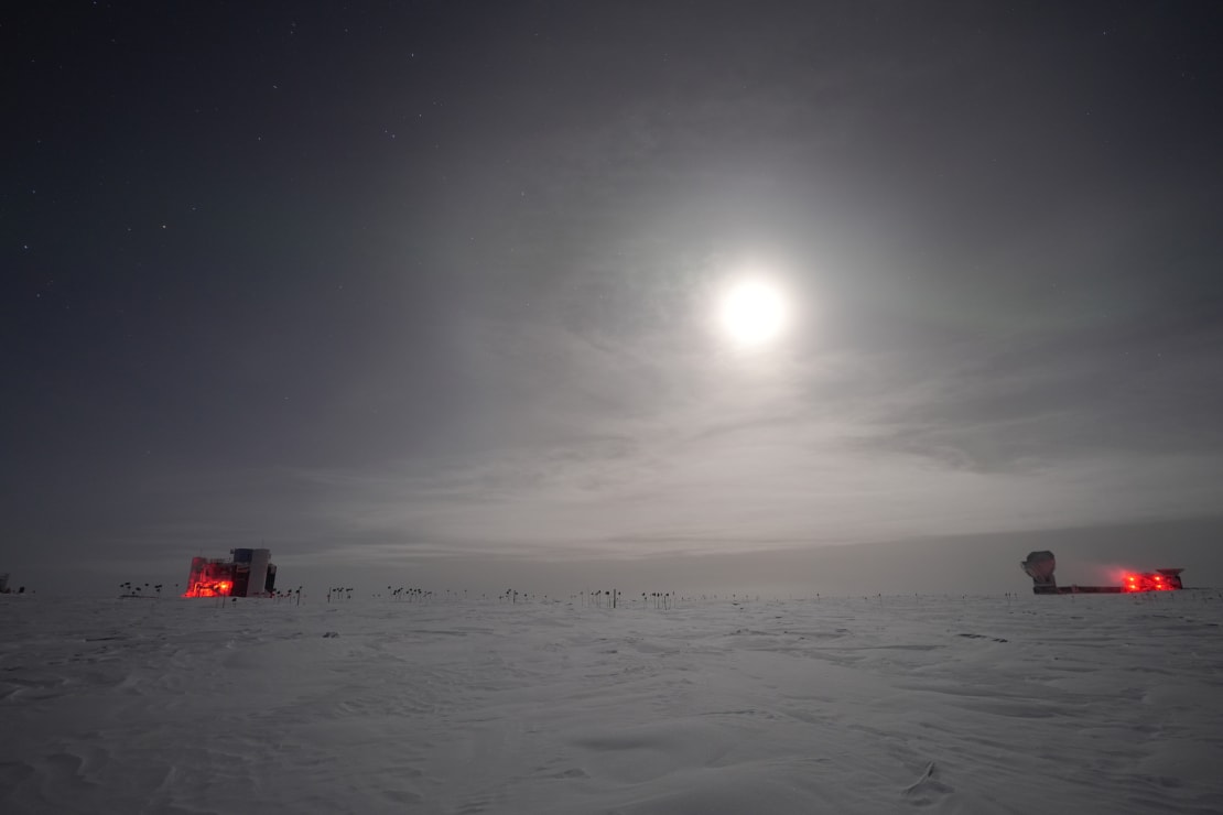 Bright moon in sky, with the IceCube Lab and South Pole Telescope in distance.