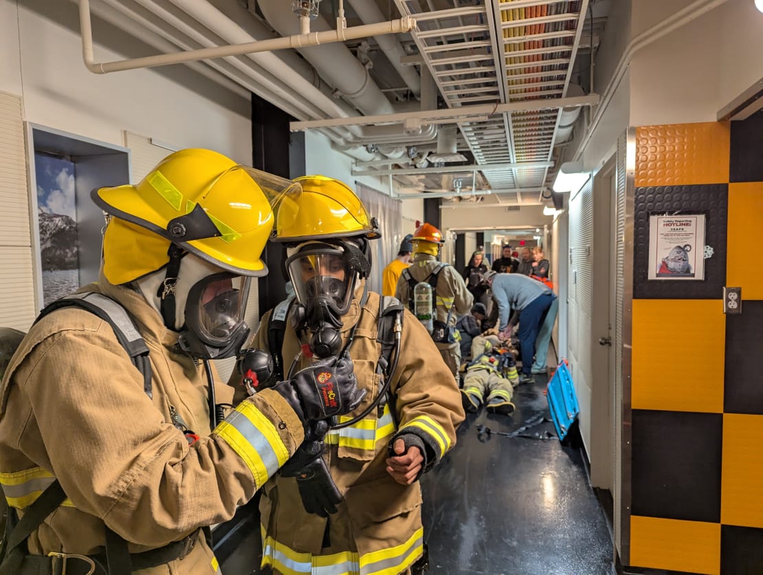 Emergency response team exercise in progress in hallway, a person in full firefighter gear on ground in background being attended to, two firefighters examining something in foreground.