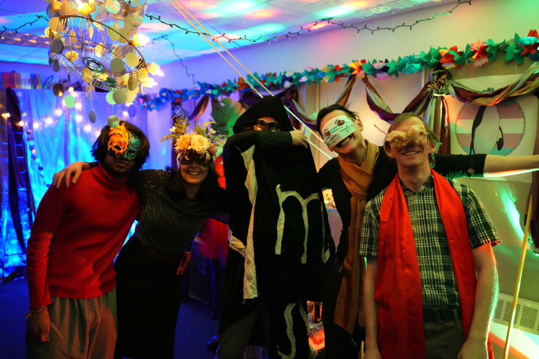 Group of five in costumes and masks, surrounded by multicolored lights and part decorations.
