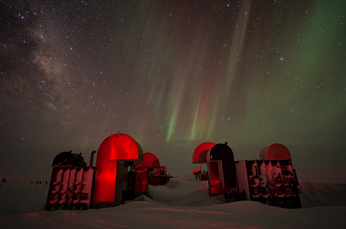 IceCube Upgrade drill camp structures reflecting red light, under clear winter sky.