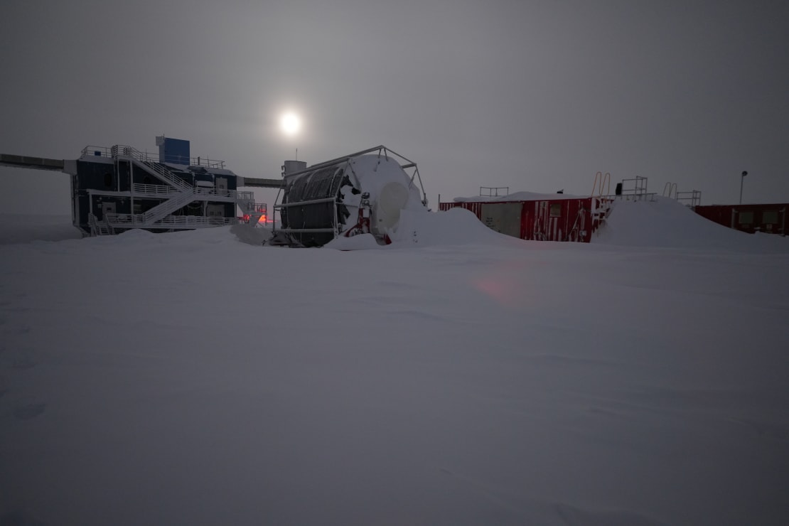 Snow-drifted hose reel and container structures, bright moon above peeking through overcast sky.