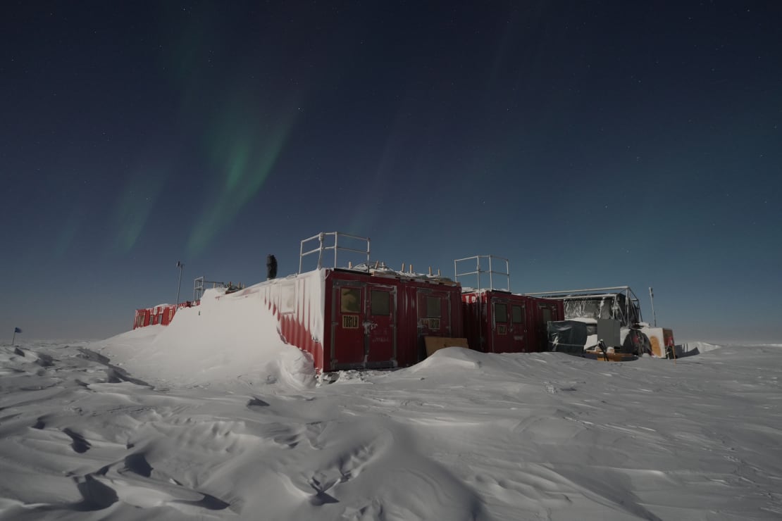 Snow drift all the way up the side of a storage container at the South Pole, light auroras overhead.