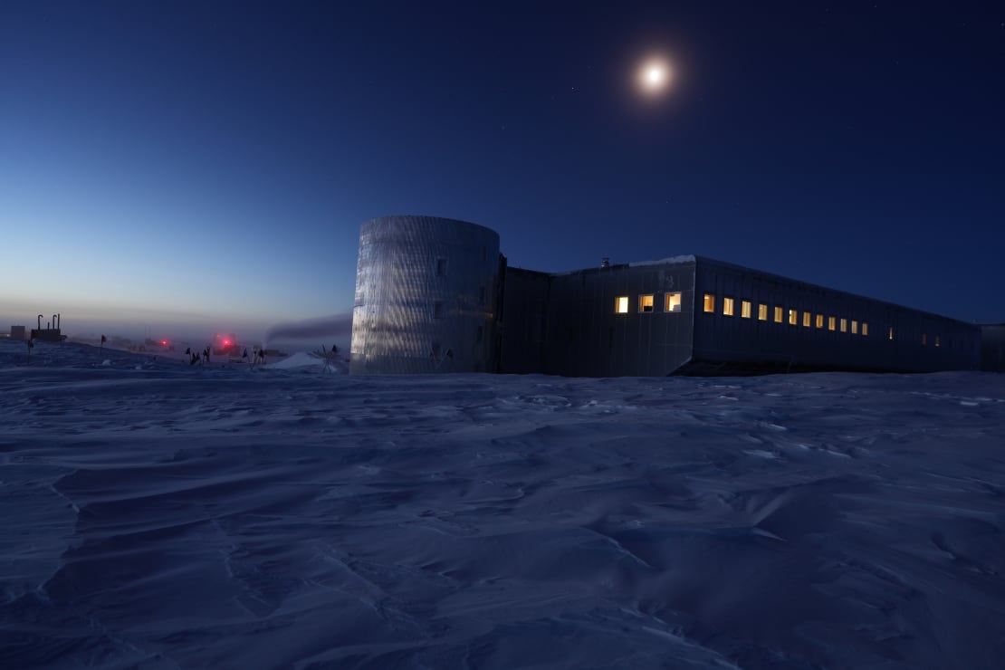 South Pole station at twilight, windows illuminated, moon overhead.