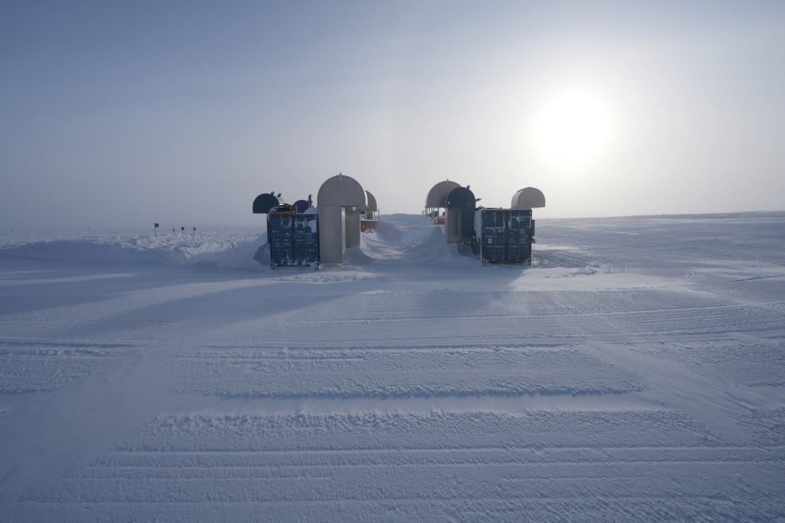 Sunny but hazy sky over the Upgrade drill camp structures showing groomed snow in the foreground.
