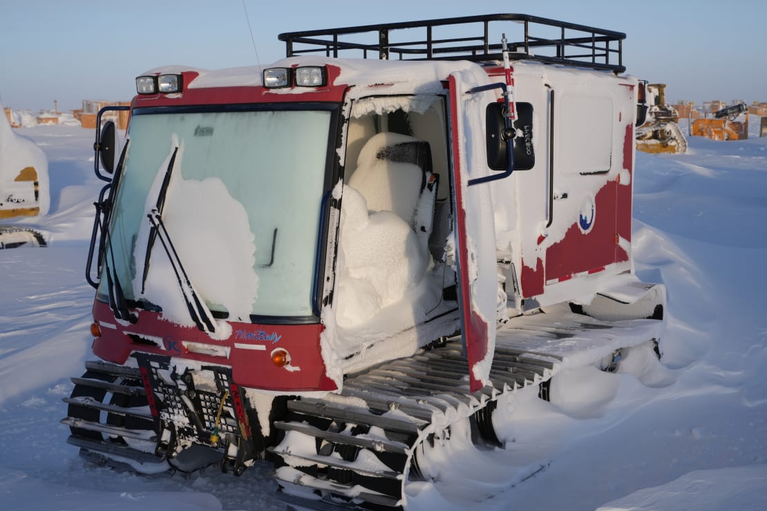 A snow-covered snow grooming vehicle stored outside, with a door ajar showing snow that has accumulated inside.