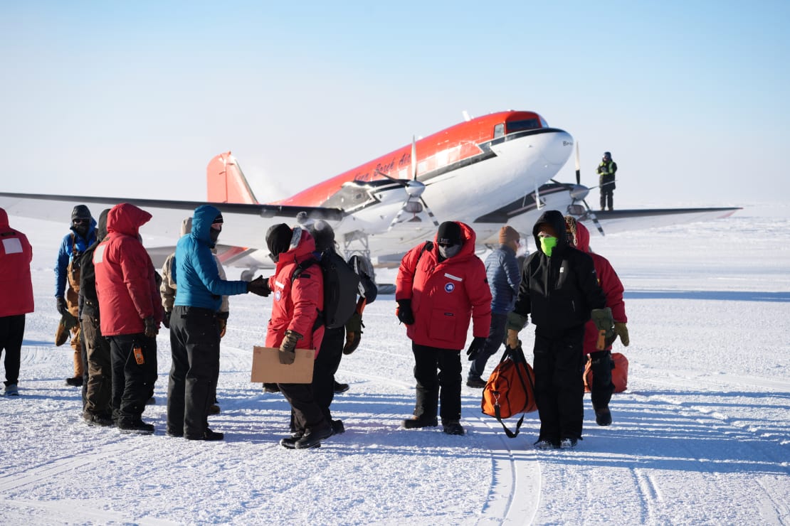 Small plane in background parked on ice, with group of arrivals in parkas in foreground.
