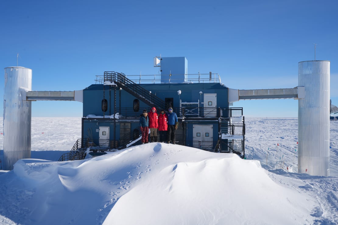 Group photo of four winterovers standing in front of the IceCube Lab with a large snowdrift in front of them.