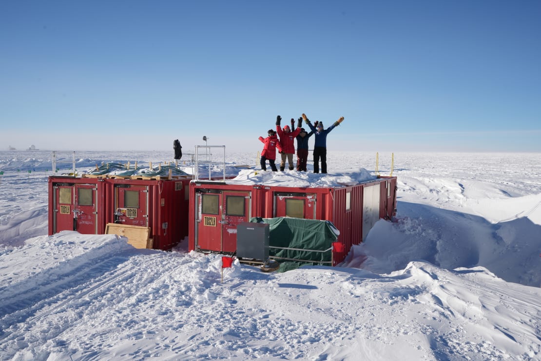 Group photo of four winterovers standing on top of one of the containers out at the IceCube Upgrade drill camp.