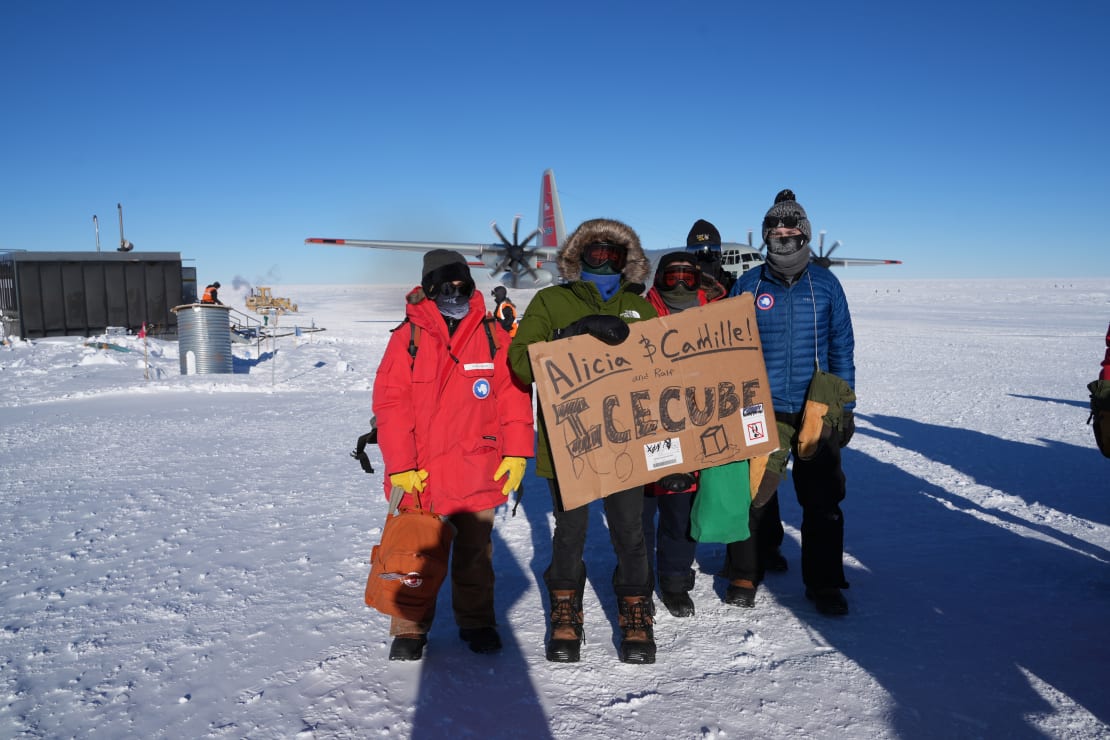 Group in parkas holding large cardboard sign out on the ice with parked plane behind them.