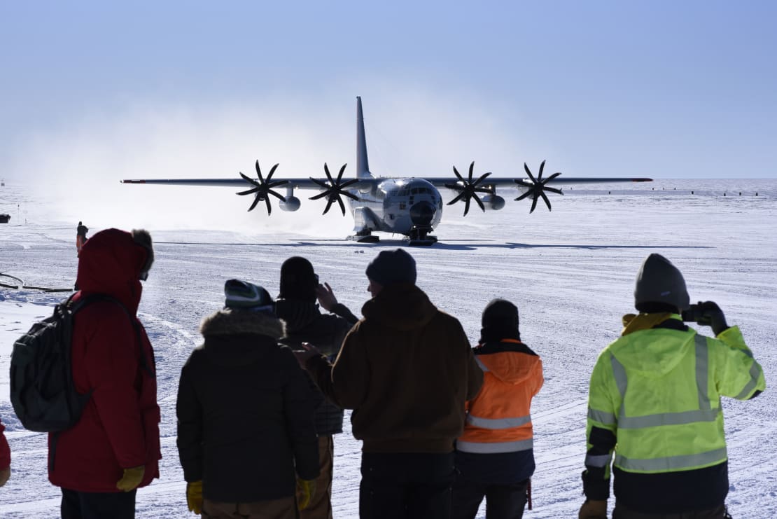 Group of people seen from behind in the foreground, large propellor plane approaching on ice in the background.