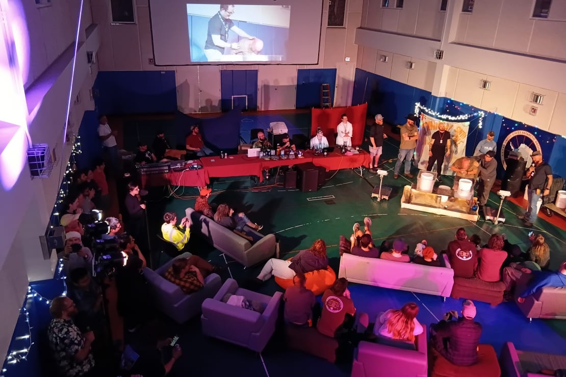 Overhead view of a gymnasium during a competition, with lots of tables, people, and chairs encircling a stage.