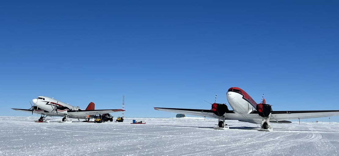 Two small planes parked side-by-side on the ice and facing forward, clear blue sky overhead.