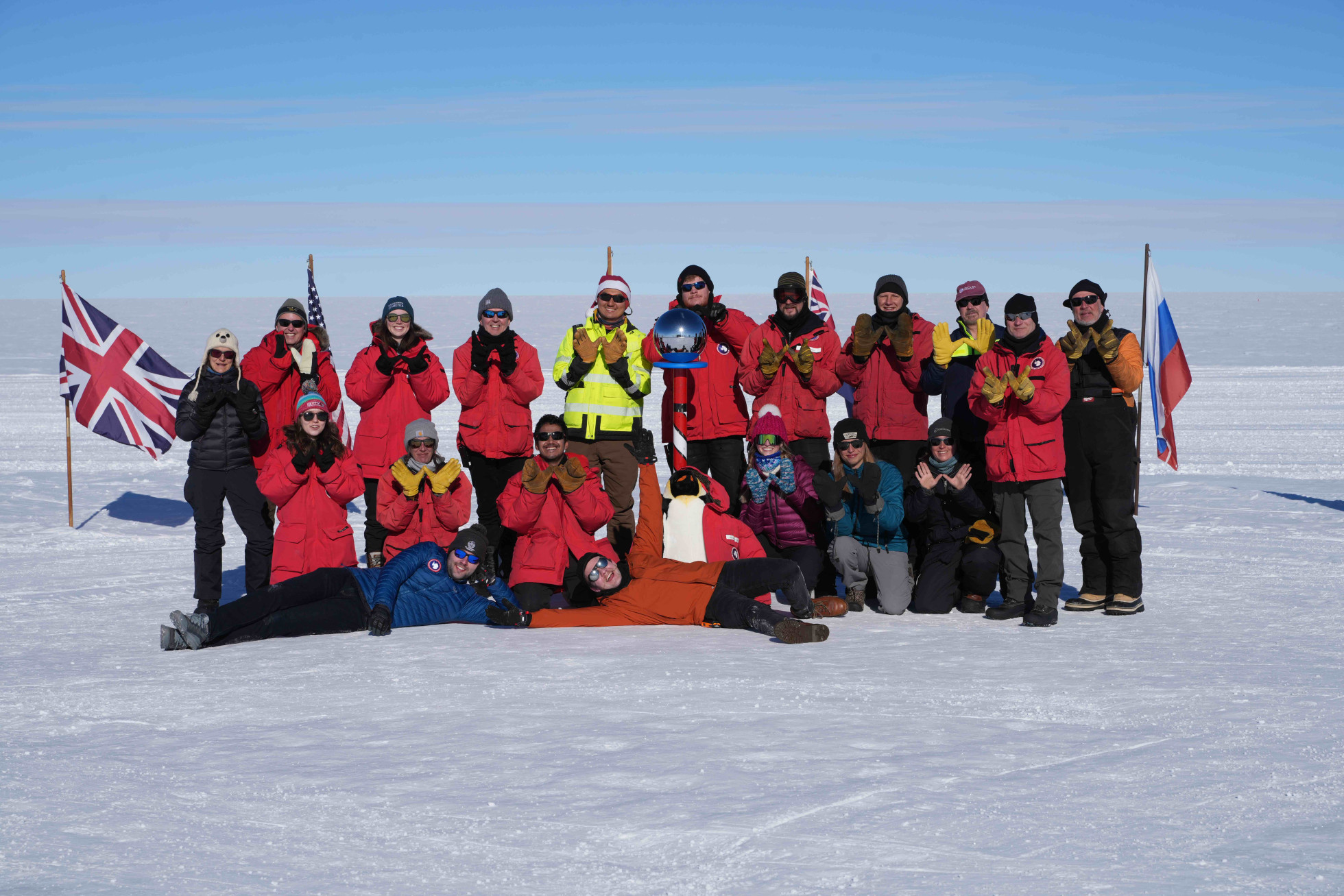 A group photo of about 20 people in parkas at the ceremonial South Pole, with raised hands "W" formations.