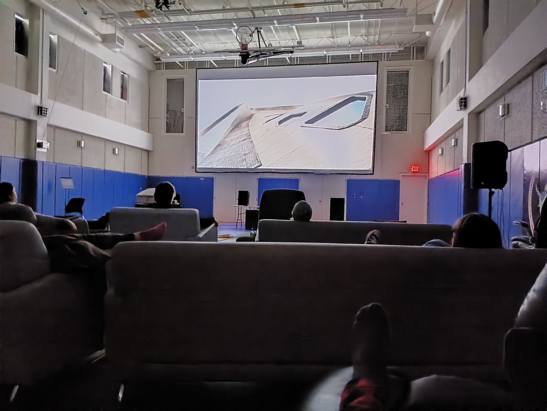 View of movie screen on wall of gym, seen from behind a few rows of occupied chairs and sofas.