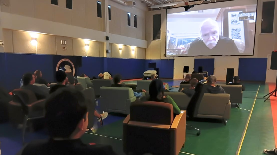 In gym set up theater style, view from behind of folks watching screen.