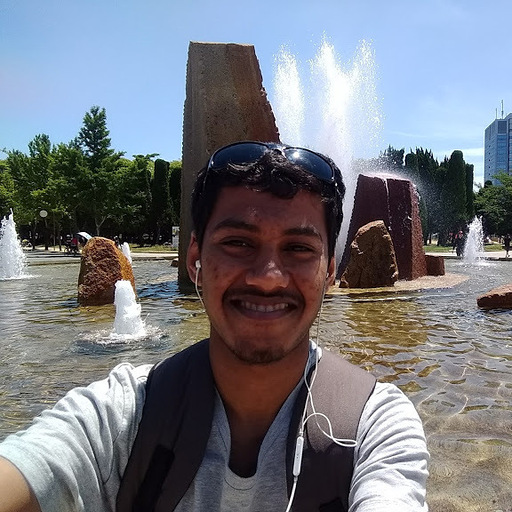 A man with sunglasses and headphones smiling in front of a water fountain.
