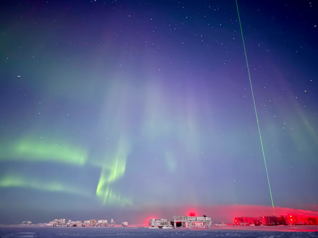 Green swirling auroras at South Pole at twilight, with a vertical laser beam off to the right.