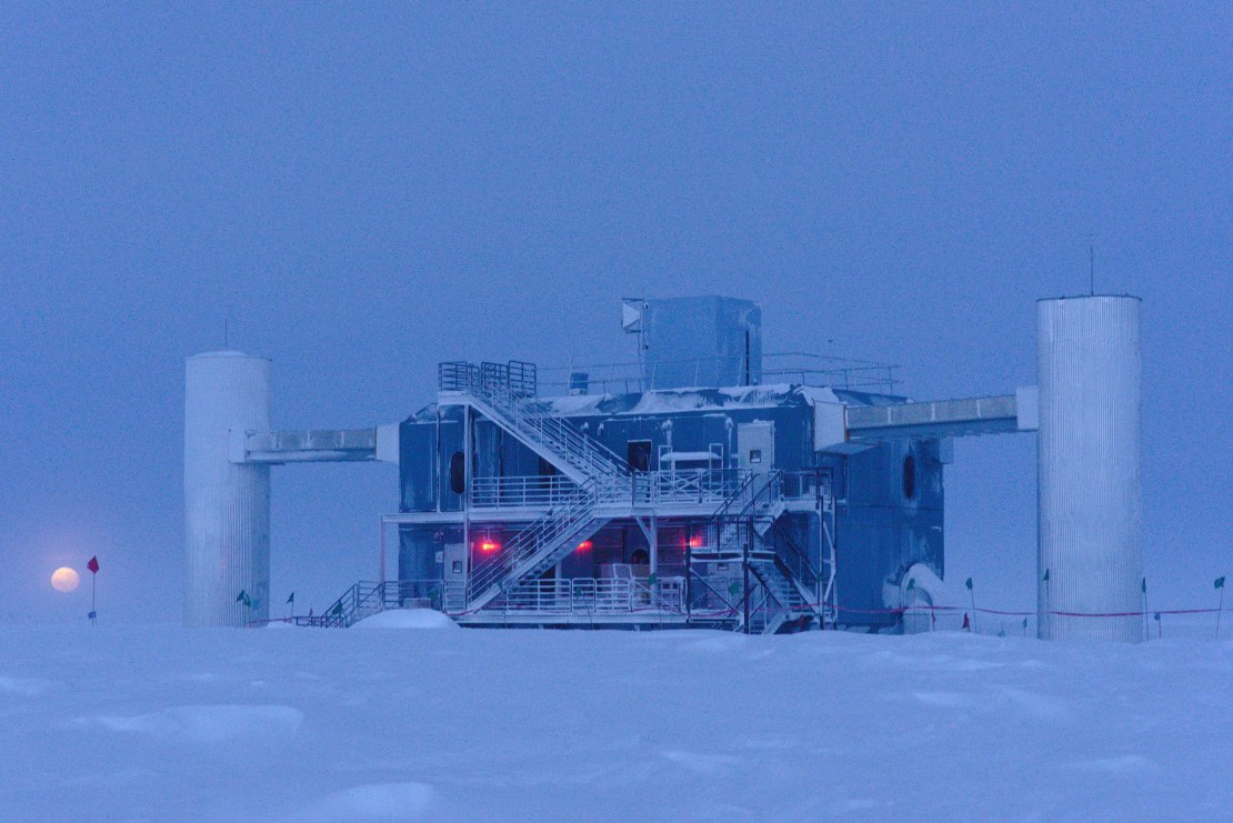 Twilight view of the IceCube Lab, with the ground and sky merged in blue and the moon low in the sky to the left.