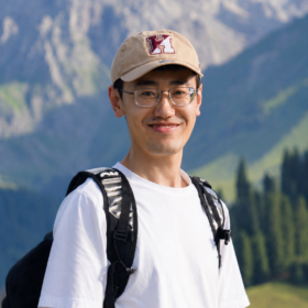 A man wearing a baseball cap and white shirt posing for a photo in front of mountains.