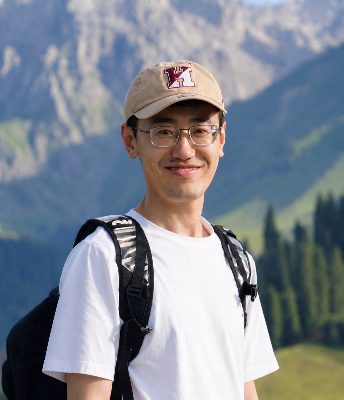 A man wearing a baseball cap and white shirt posing for a photo in front of mountains.