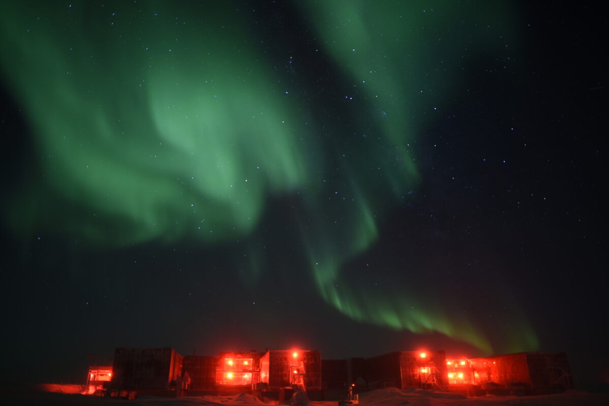 Swirling green auroras in dark sky above cluster of red lights on building exteriors.