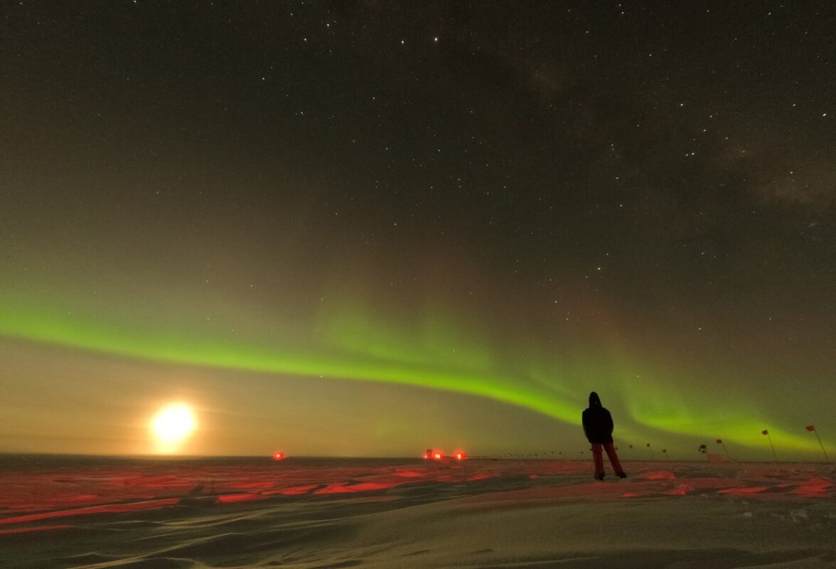 Bright moon low on horizon with a strong line of green auroras crossing the sky above it.
