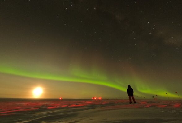 Bright moon low on horizon with a strong line of green auroras crossing the sky above it.