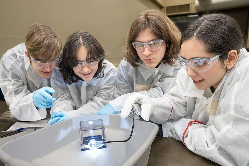 A group of students in personal protective equipment huddled around a cloud chamber.