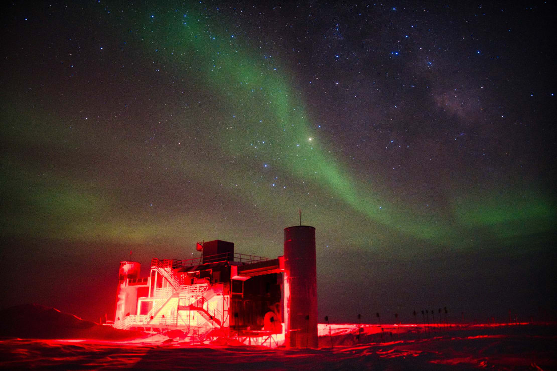 Three-quarter view of the IceCube Lab lit in red, with stars and auroras in the dark sky above.