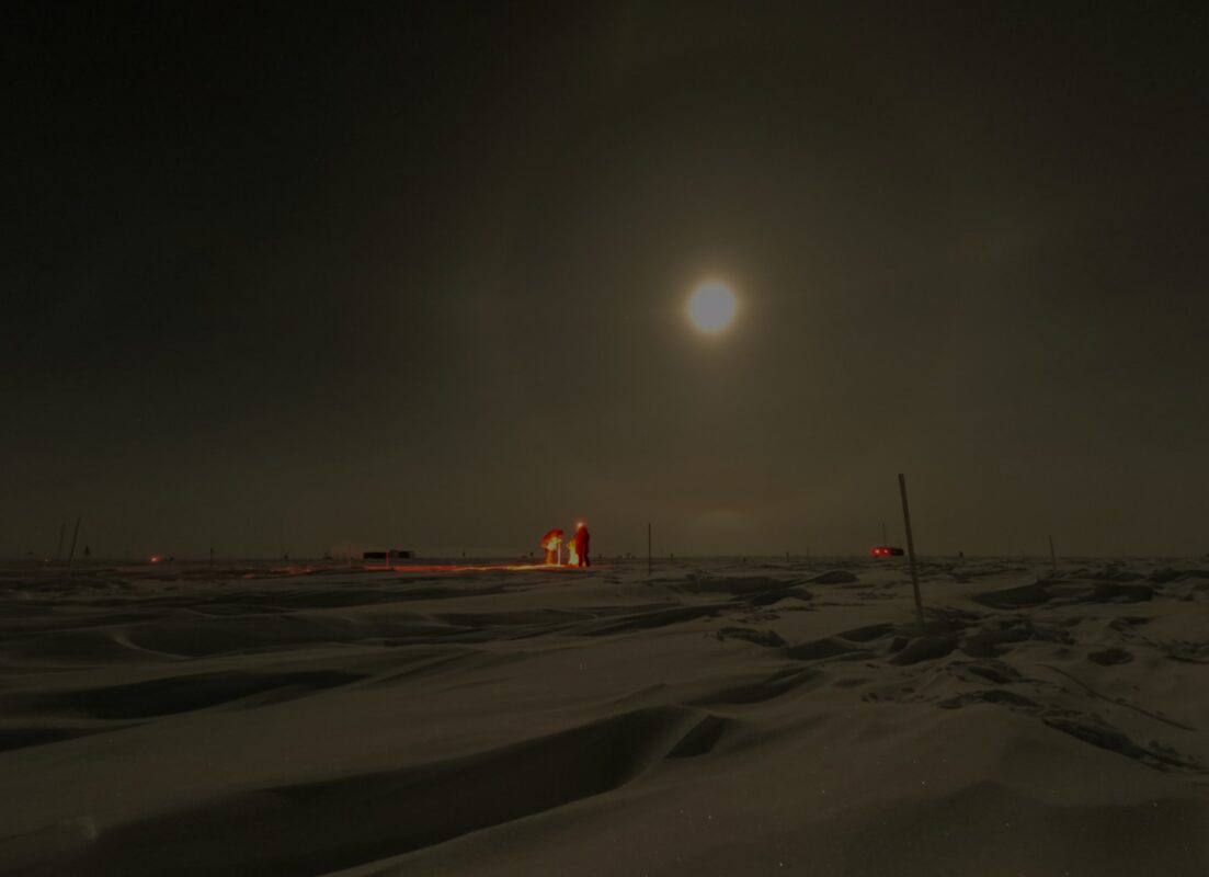 Dark photo with full moon in the sky above two people working under lights in the distance.