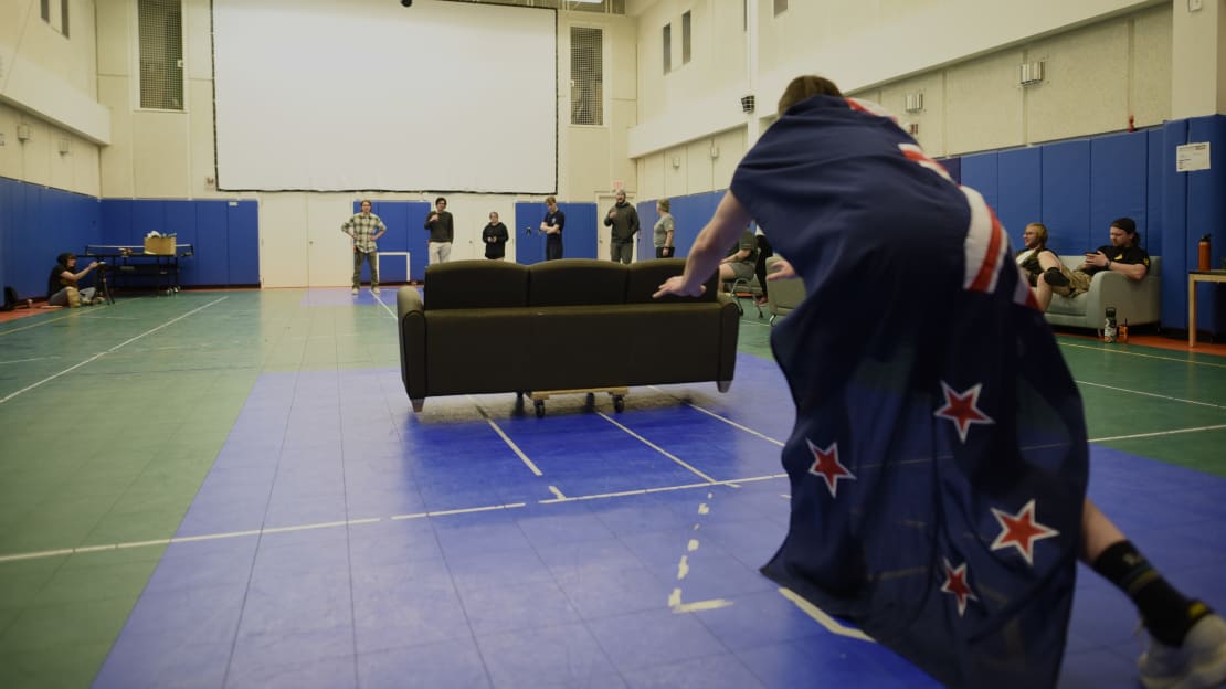 A person seen from behind as they push a sofa in place of a curling stone in the middle of the gym.