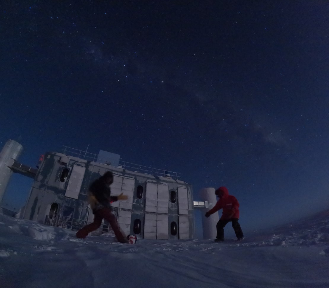 Two people in parkas kicking a soccer ball in front of the IceCube Lab, in the dark under a starry sky.