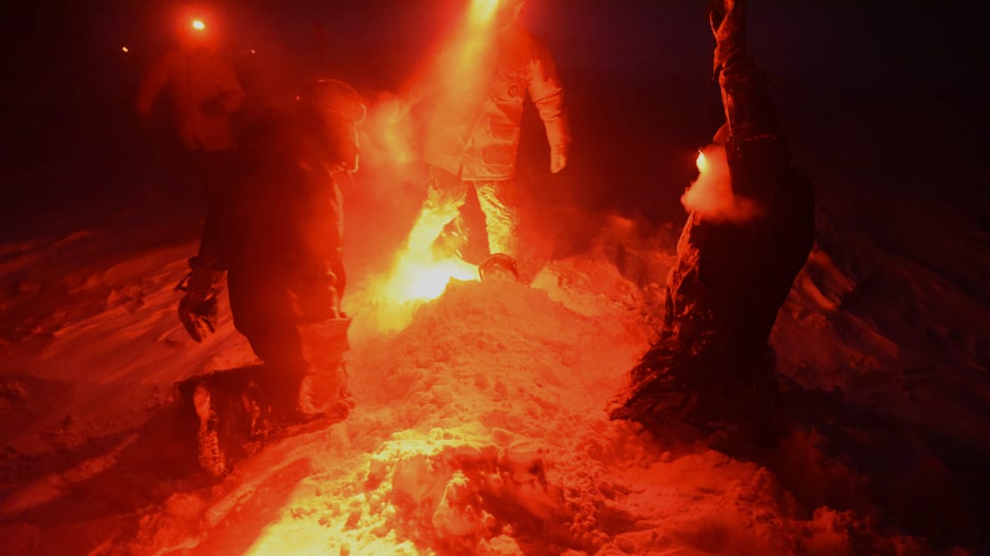 Person lying down buried in snow, as one might be on a beach, but in the Sout hPole darkness under headlamps.