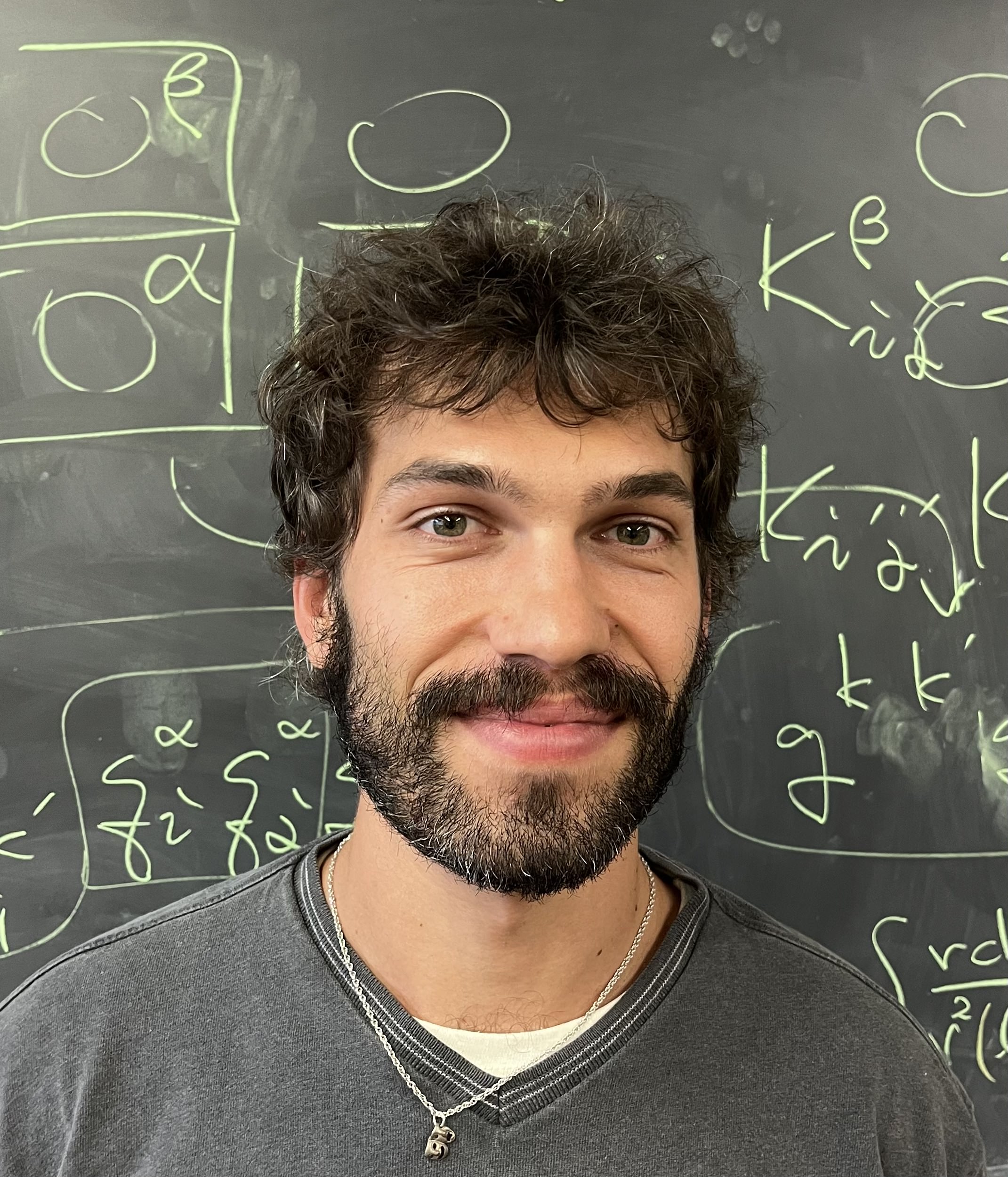 A headshot of a man with curly brown hair in front of a chalkboard.
