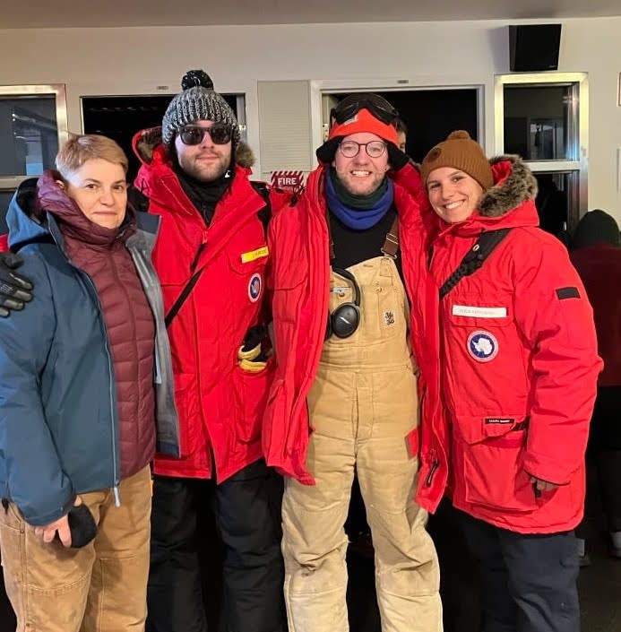 Four people wearing big winter coats pose for a photo at the South Pole Station.